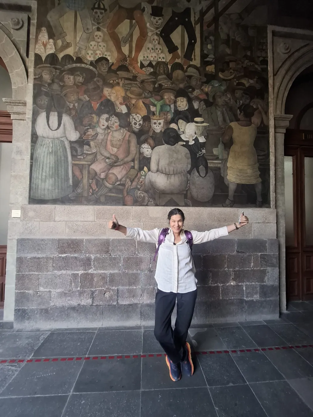 Mely with arms outstretched beneath a historic Mexican mural