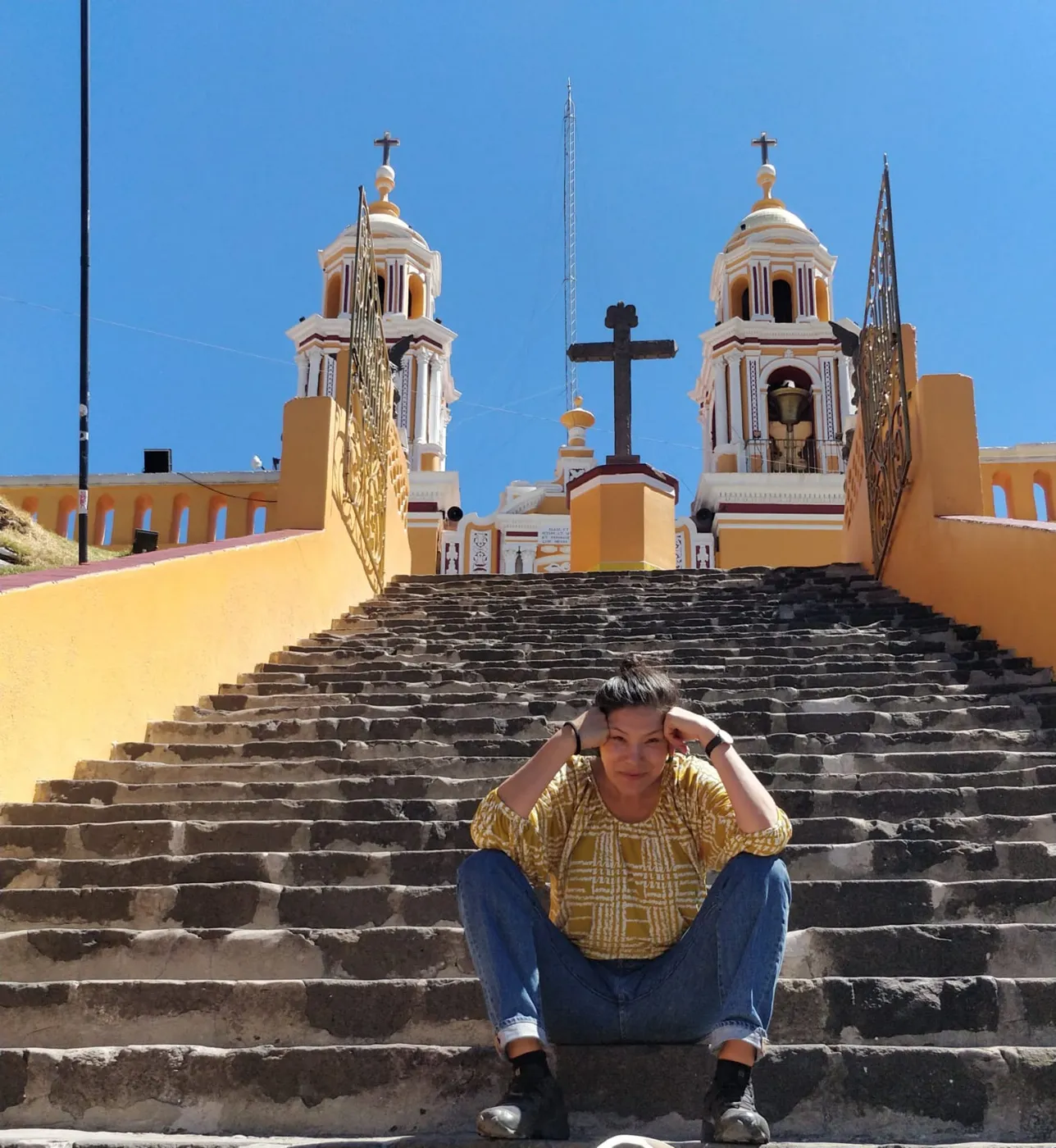 Mely sitting on the stone steps of a yellow church in Cholula under a bright blue sky
